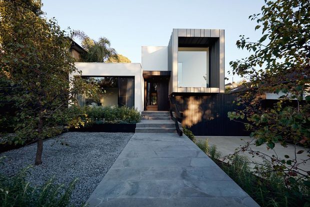 A street-facing view of a contemporary two-storey residence with crisp rectilinear volumes, white and dark cladding, recessed glazing, and a broad concrete path leading to the entry. The house is surrounded by mature landscaped planting and a gravel garden, with strong contrast between the light upper volume and dark lower facade.