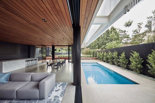 Contemporary open-plan indoor-outdoor living space with a modern kitchen, dining area and lounge opening directly onto a narrow rectangular swimming pool. The interior features a warm timber-lined ceiling, dark structural columns and frames, polished light concrete flooring, minimalist grey furnishings, and extensive glazed openings. Outside, the pool is bordered by light paving, dense screening plants and a dark vertical slat fence.