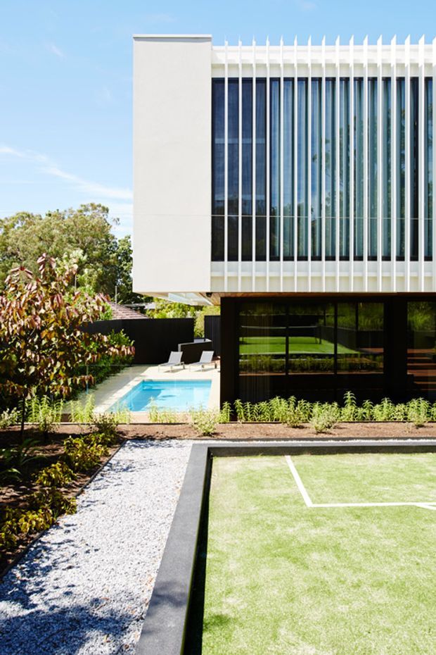 Contemporary multi-level building with a crisp white rendered upper volume, extensive dark-framed glazing, and a distinctive vertical white fin facade. The landscaped grounds include a swimming pool, gravel path, planted garden beds, and a small outdoor sports court in the foreground.