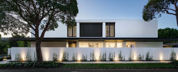 A contemporary two-storey residential facade viewed from the street at dusk. The minimalist white-rendered upper volume features a large dark vertical-panel opening and narrow timber-toned screened windows, while the lower level is set behind a tall white boundary wall with warm architectural lighting and slender planted greenery. Mature street trees frame the composition.