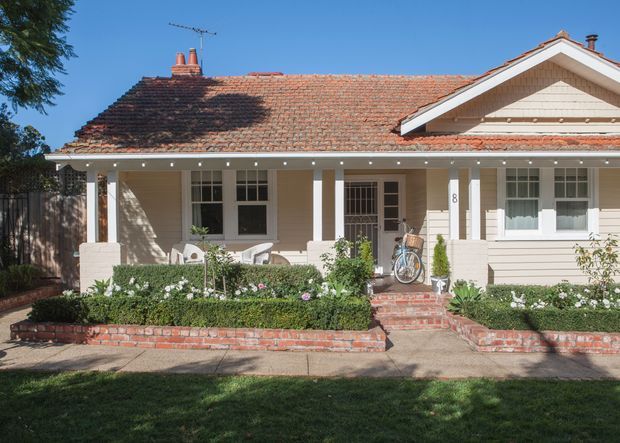 Front exterior view of a single-storey traditional bungalow with a broad terracotta tiled pitched roof, cream weatherboard cladding, white timber trims, a covered verandah, and a landscaped front garden with brick retaining walls and steps.
