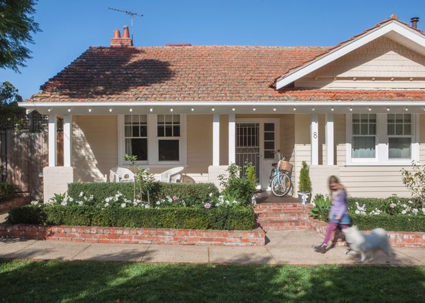 Street-facing single-storey character home with a broad tiled gable roof, pale weatherboard-style exterior, covered front verandah supported by square white columns, brick steps and low brick garden edging. The front garden is landscaped with clipped hedges and flowering plants, while the facade includes traditional multi-pane windows and brick chimneys.