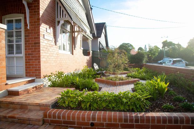 Sunlit front garden and entry courtyard beside a brick residential house, featuring terracotta brick retaining walls, paved steps and paths, raised garden beds, and established planting. The facade includes traditional multi-pane windows, decorative timber detailing, and a pitched roof.