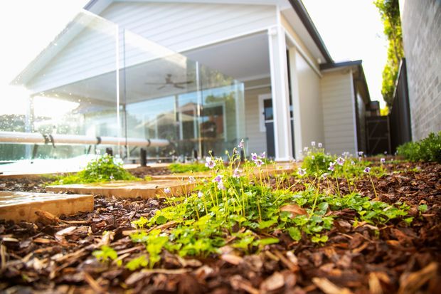 Bright exterior view of a contemporary white-clad home extension or sunroom with a pitched gable roof, extensive floor-to-ceiling glazing, and a small landscaped garden bed in the foreground. A paved terrace and outdoor seating are visible through the glass.