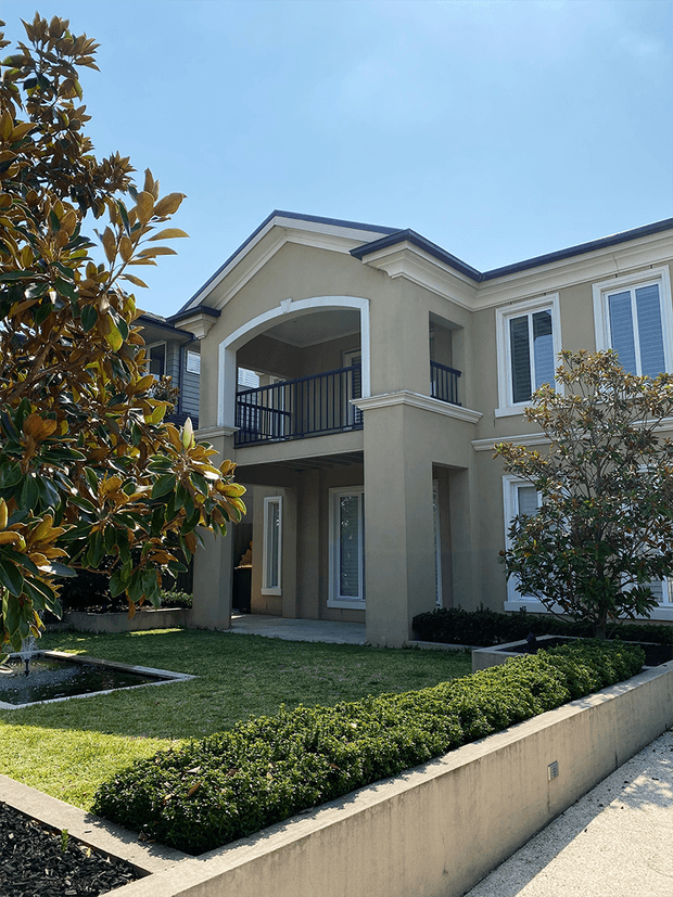 Two-storey residential exterior with a pale rendered facade, dark pitched roof, prominent arched balcony opening, and black metal balcony balustrade. The building is framed by manicured hedges, lawn, raised concrete garden beds, and established trees in a formal landscaped setting.