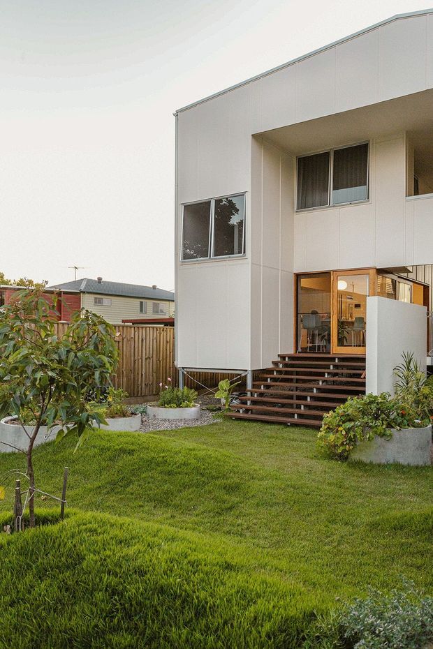 A contemporary two-storey residential house viewed from the landscaped front garden. The facade is clad in pale, smooth panelled material with large rectangular windows and a recessed upper-level balcony or entry void. Timber-framed glazing opens onto a broad timber staircase and deck. The garden includes manicured lawn, raised concrete planters, gravel, shrubs, and a timber boundary fence, with neighbouring suburban homes visible beyond.