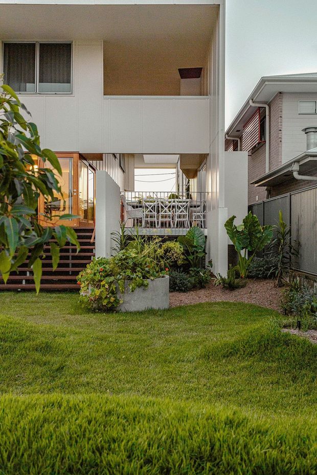 Front exterior view of a contemporary elevated residential house with white vertical-panel cladding, an upper covered balcony, timber-framed glazed entry, open stair access, and planted garden beds across a lawn. The house is closely bordered by neighbouring dwellings and fencing.