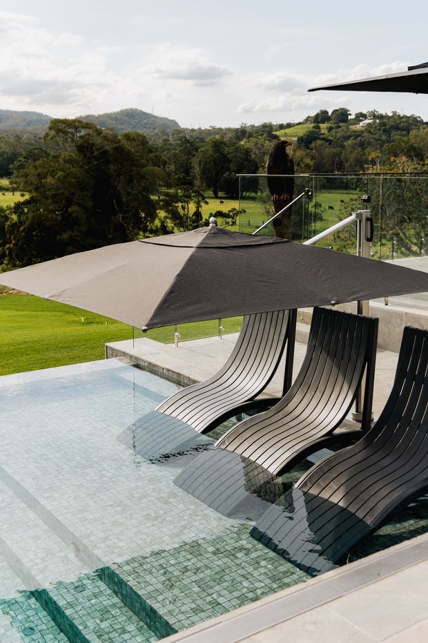 An outdoor pool terrace overlooking a lush rural, hilly landscape. The shallow tiled pool contains sculptural black lounge chairs partially submerged in the water, shaded by large cantilevered umbrellas. A frameless glass balustrade and concrete paving define the contemporary terrace.