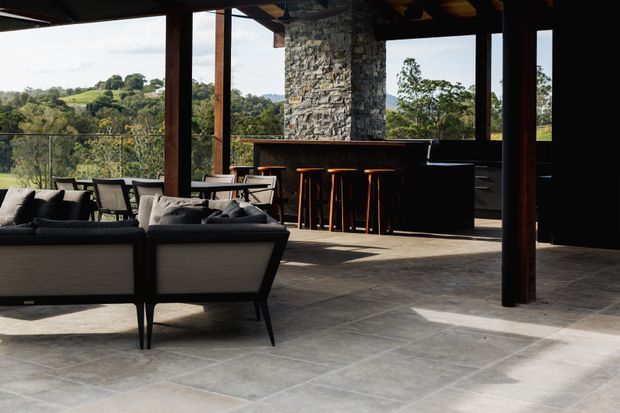 A spacious, open-plan indoor-outdoor living area with expansive glazing overlooking a green rural landscape. The room features a dark timber structural frame, a rough natural-stone column, large grey stone floor tiles, a dark kitchen or bar with timber stools, and a contemporary lounge seating arrangement.