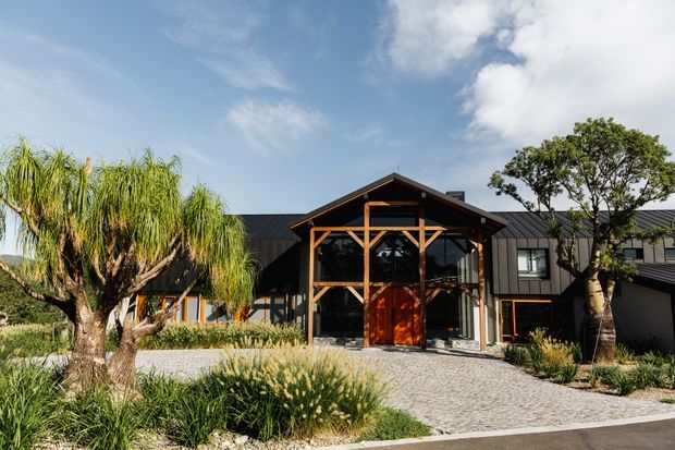 Exterior view of a contemporary dark-clad building with a prominent timber-framed, glazed central entry pavilion and large timber entrance door. A cobbled driveway and landscaped garden with ornamental grasses and sculptural trees lead to the entrance. The roof form is predominantly pitched with visible timber structural framing.