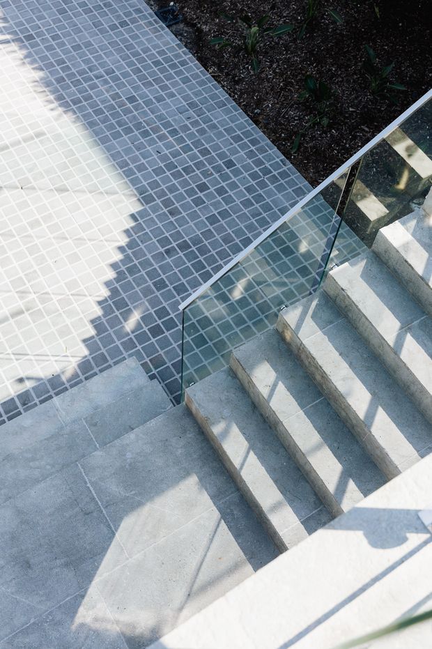 Exterior architectural detail showing a concrete stairway descending beside a large paved terrace or poolside area. The stairs have a clear glass balustrade with slim metal supports, and the adjacent surface is finished in small square grey mosaic tiles. Dark planted garden beds border the tiled area.