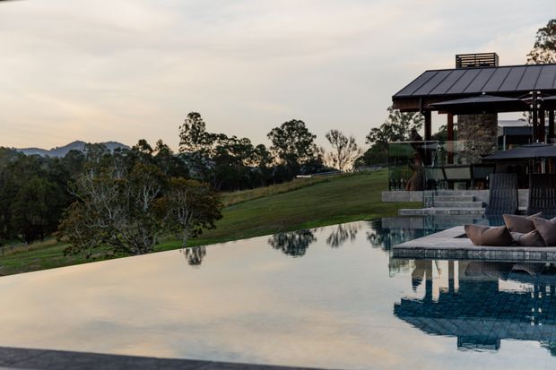 A landscaped hillside property at dusk with a large reflecting swimming pool in the foreground. The pool overlooks rolling rural terrain and trees, while a partially visible open-sided pavilion with a dark pitched roof, stone-clad wall, glass balustrade, and outdoor lounge seating occupies the right side.