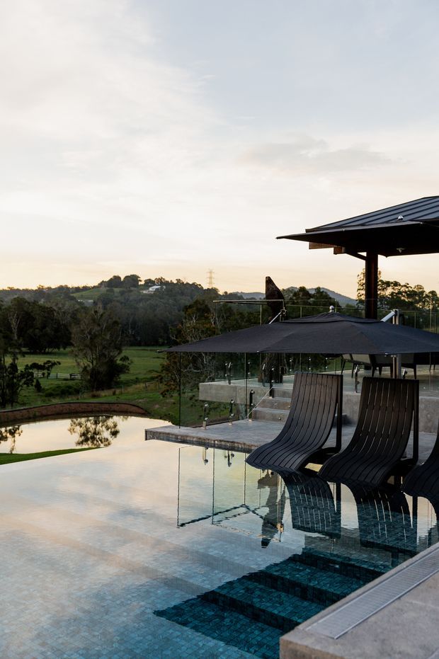 A contemporary outdoor pool terrace overlooking a green rural landscape. The infinity-style pool has dark blue mosaic tile steps and submerged sculptural loungers, with large black umbrellas, glass balustrades, and a covered pavilion structure. The scene is softly lit at sunset and includes a secondary pond and rolling wooded terrain in the distance.