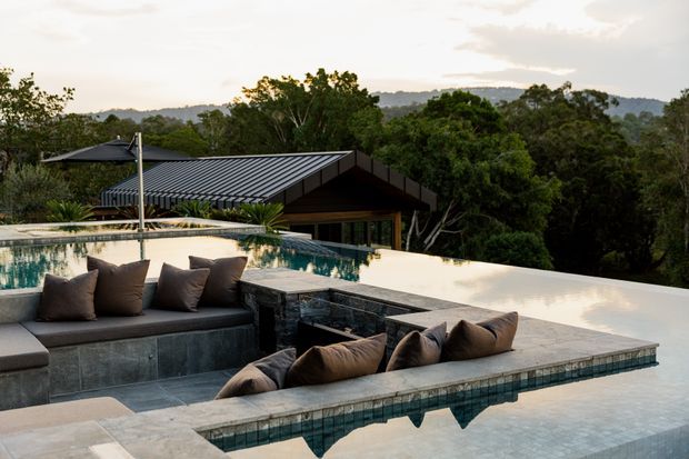 A sophisticated outdoor pool and lounge area overlooking a wooded, rolling landscape. The image features a long reflecting pool with integrated sunken seating, cushioned built-in lounge benches, pale stone paving, and a dark metal pitched-roof house nestled among trees in the background.