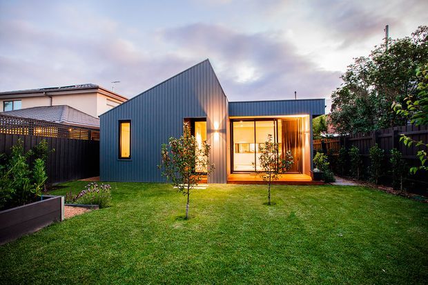 A compact modern backyard dwelling with a dark vertical metal-clad gabled volume connected to a low flat-roofed glazed extension. Warm interior lighting illuminates the large window wall, while a manicured lawn, raised garden bed, timber fencing, and established planting surround the structure. A neighbouring suburban house is visible beyond the fence.