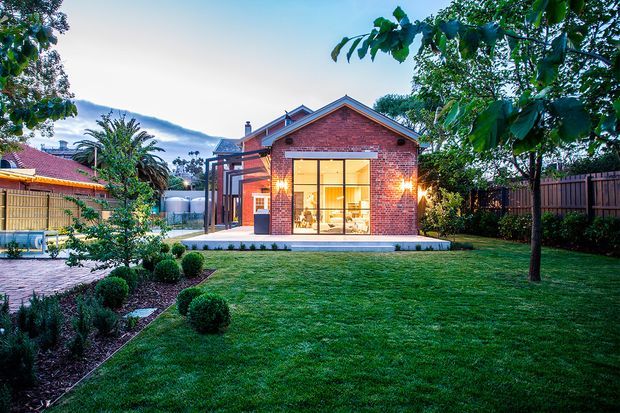 A single-storey red-brick building set within a landscaped suburban garden, photographed at dusk. The gabled facade features a large black-framed glazed opening with warm interior lighting, flanked by exterior wall lights. A paved path, clipped shrubs, lawn, mature trees, timber fencing, and neighbouring rooflines surround the structure.