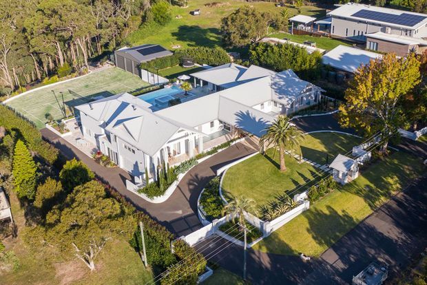 Aerial view of a large contemporary residential property with multiple interconnected white-roofed wings, landscaped gardens, curved driveways, a swimming pool, tennis court, detached outbuildings, and surrounding mature vegetation. The home sits in a spacious suburban or semi-rural setting.