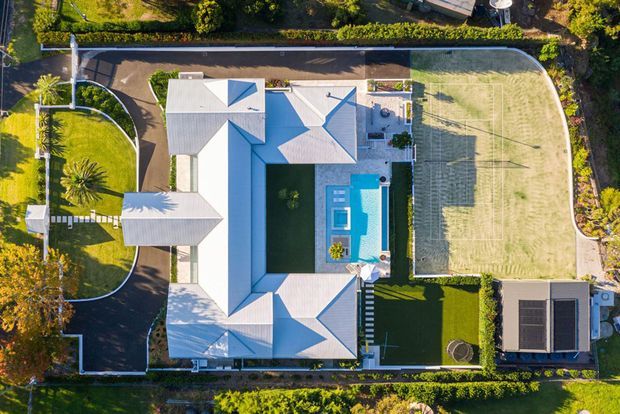 Aerial top-down view of a large residential property with multiple light-colored pitched roof volumes arranged around a central landscaped courtyard and rectangular swimming pool. The site includes manicured lawns, curved paved paths and driveways, dense perimeter planting, a fenced multi-purpose sports court, and a separate covered structure with solar panels.