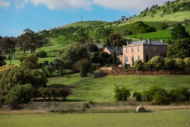 A stately two-storey brick country house set within expansive green rolling farmland, with mature trees, landscaped grounds, stone retaining walls, and surrounding cultivated hills. The building has a symmetrical traditional facade, pitched slate-coloured roof, sash-style windows, and a small central entrance porch. Bright rural landscape photography with a clear blue sky.