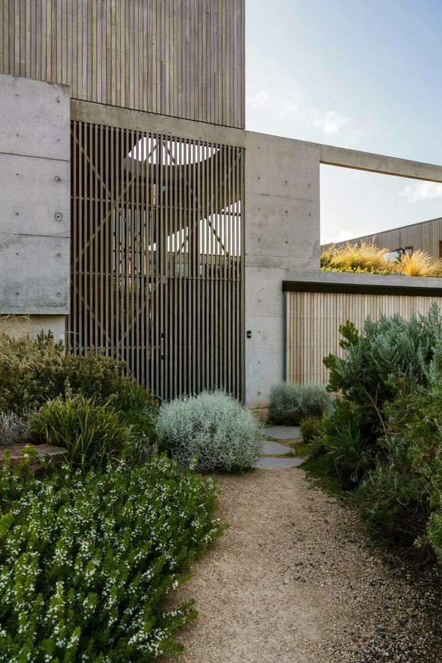 A contemporary residential exterior viewed from a landscaped garden path. The façade combines smooth precast or poured concrete walls, vertical timber cladding, and a tall vertical-slatted screen enclosing an entry or carport-like opening. Gravel paving, stepping stones, and dense drought-tolerant planting frame the approach. A partially visible low roof and linear concrete beam reinforce the minimalist geometric composition.