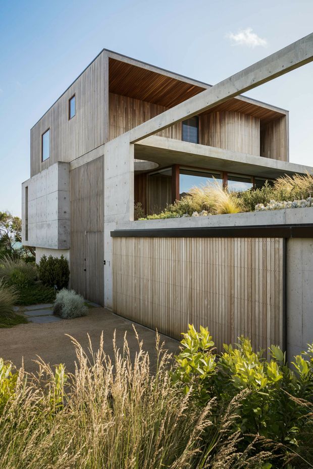 A contemporary multi-level residential building viewed from the landscaped exterior. The facade combines board-formed concrete elements with vertical timber cladding, deep recessed balconies and warm timber soffits. A concrete-framed upper terrace contains grasses and low planting, while a timber-clad garage door and landscaped path occupy the foreground.