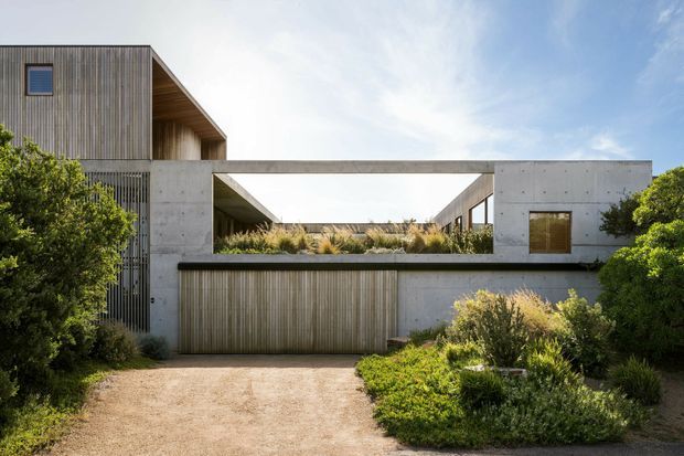 Contemporary residential facade with a strong geometric composition, combining raw concrete walls, vertical timber screening, and a recessed open courtyard or terrace framed by a rectangular concrete opening. A timber-clad upper volume rises on the left, while dense coastal-style planting surrounds the gravel driveway and integrated garage door.