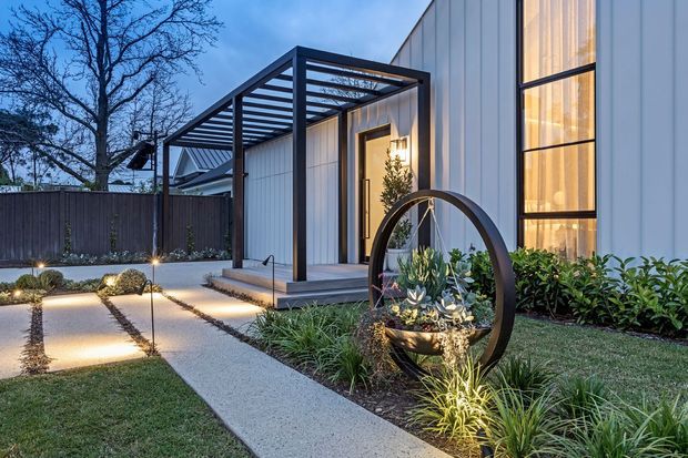 Contemporary residential front entry at dusk, featuring a white vertically panelled facade, tall illuminated glazing, a dark steel pergola over the porch, concrete garden paths, and carefully landscaped planting beds with a circular sculptural planter.
