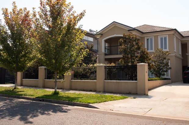 A suburban two-storey residential house viewed from the street, featuring a warm beige rendered facade, tiled pitched roof, gabled upper-level balcony, white-framed windows with shutters, and a black metal fence set on a low rendered wall. A paved driveway and landscaped verge are visible in front.