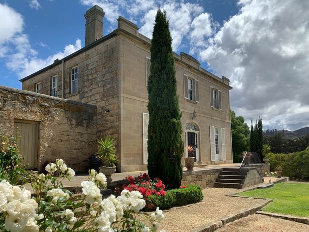 A stately two-storey stone residence set within a formal landscaped garden. The pale ashlar masonry facade features tall shuttered windows, classical trim, a decorative fanlight above the entrance, substantial chimneys, and a cypress-lined gravel forecourt with flowering rose beds and stone steps.
