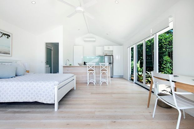Bright open-plan coastal-style apartment interior combining a bedroom and compact kitchen-living area. The space features pale timber-look flooring, white cabinetry, a light island with woven bar stools, a white upholstered bed, a simple dining/work table, extensive full-height glazed doors, and lush greenery outside. White walls, recessed lighting, and a ceiling fan create an airy, relaxed atmosphere.