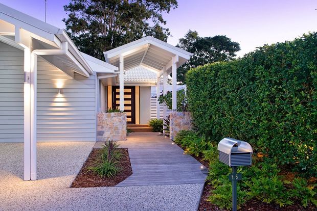 A landscaped residential front entry at dusk, featuring a light grey horizontal-clad garage and house facade, a covered gabled porch with white posts and exposed rafters, a timber-look walkway, stone-clad low walls, dense hedging, and subtle exterior lighting.