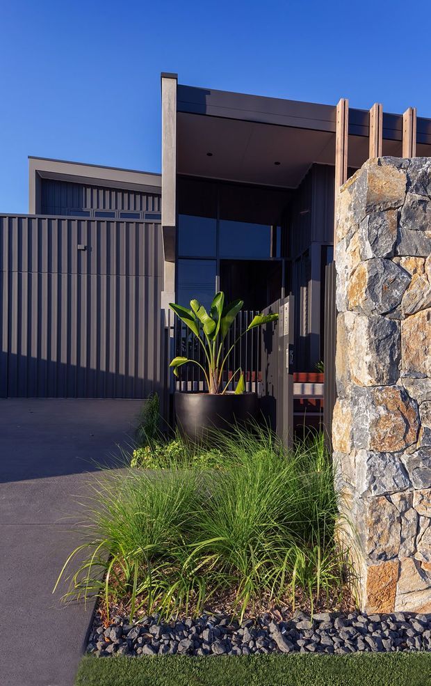 A contemporary dark-clad residential entrance viewed from the front driveway. The facade features vertical corrugated metal cladding, large dark-framed glazing, a broad covered entry canopy, timber posts and detailing, and a substantial natural stone boundary wall. Native-style grasses, black gravel and a large potted tropical plant provide landscaping around the entry.