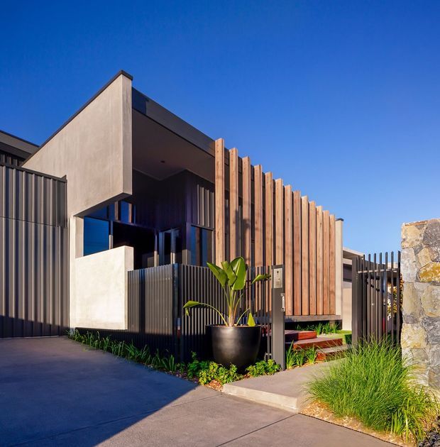 Contemporary residential facade viewed from the street, featuring a geometric concrete entry volume, dark corrugated metal fencing and cladding, vertical timber screening, glazing, paved driveway and landscaped planting. A low stone wall frames the right edge of the property under a clear blue sky.