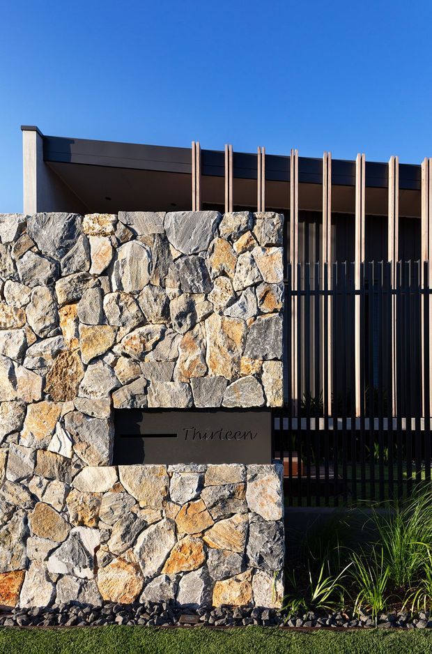 A modern residential entrance viewed from outside, featuring a substantial irregular natural-stone masonry wall with a dark address plaque, vertical timber screening, black metal fencing, and a flat-roofed facade under a clear blue sky. Ornamental grasses and gravel landscaping line the base.