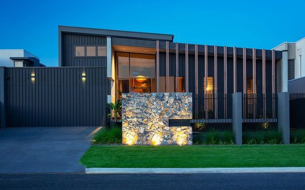 Contemporary multi-level residential facade photographed at dusk, featuring dark vertical metal cladding, strong geometric massing, recessed warm-lit entry glazing, vertical timber or metal screening, and a prominent illuminated natural-stone feature wall at the front boundary. A neat lawn, driveway, boundary fencing, and neighboring modern volumes complete the streetscape.