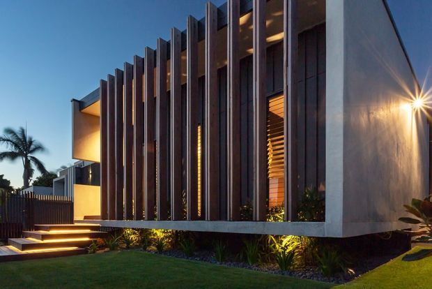 Dusk exterior view of a contemporary multi-level residence with a strongly articulated facade of tall dark vertical fins, warm recessed lighting, timber-toned louvres, and smooth pale rendered walls. Stepped illuminated entry stairs, landscaped planting, and a lawn frame the building.