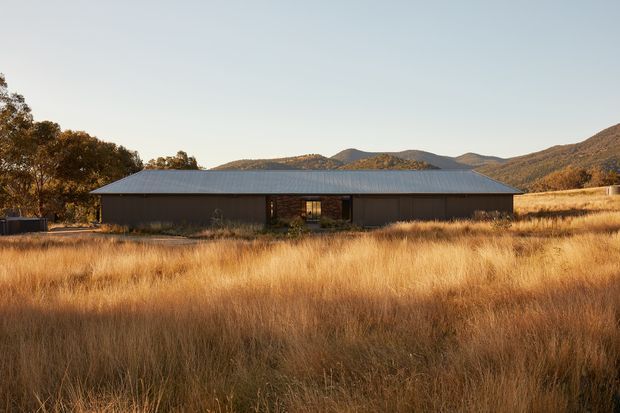 A low, single-storey rural dwelling set within tall golden grass, with a long dark metal-clad facade, broad shallow pitched metal roof, and a recessed central entry or glazed opening. Tree-covered hills and a mountainous landscape form the background. The image is an exterior architectural photograph in warm late-afternoon light.