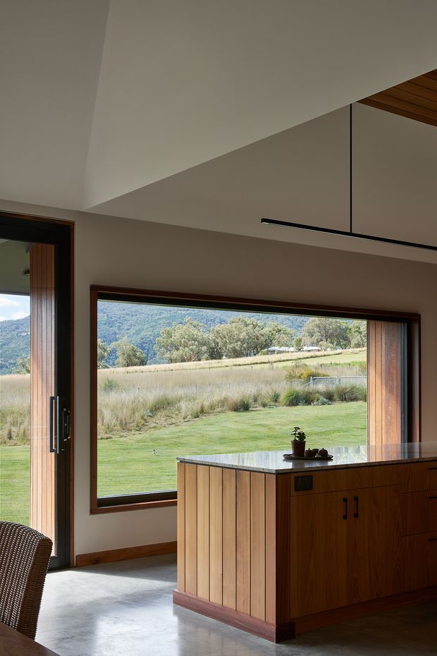 A warm, modern kitchen interior with a timber island and dark stone benchtop, positioned beside large timber-framed glazing overlooking a landscaped rural hillside. The room has a polished concrete floor, high angled plasterboard ceiling, timber ceiling lining at the upper right, and a slim linear pendant light. The open connection to the outdoor landscape gives the space an airy, contemporary character.
