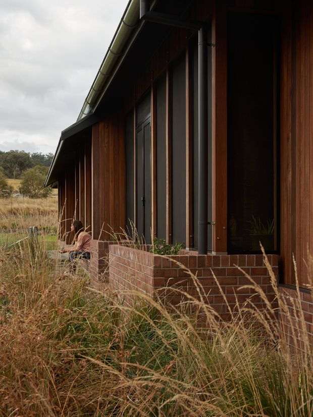 A close exterior view of a long, single-storey building set in a grassy rural landscape. The facade combines dark vertical timber cladding, tall dark-framed glazing and doors, exposed brickwork at the lower wall, and a shallow pitched roof with metal gutters. Native grasses surround the building, with a seated person near the entrance.