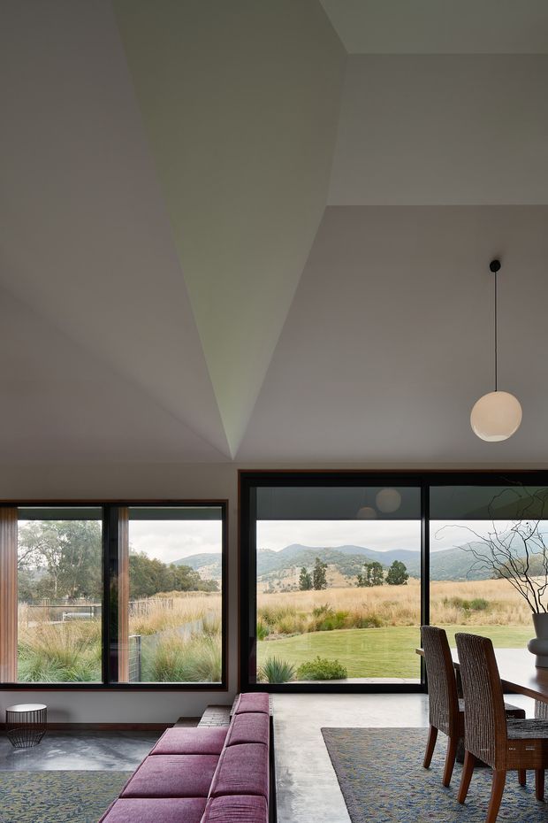 A refined open-plan living and dining area with expansive dark-framed floor-to-ceiling glazing overlooking a landscaped lawn, dry grassland, and rolling hills. The room has a sculptural angular white ceiling, dark timber window surrounds, a long deep-purple upholstered bench sofa, patterned rugs, a timber dining table with woven dining chairs, and a suspended spherical pendant light. The interior is softly lit and connects strongly to the natural landscape outside.