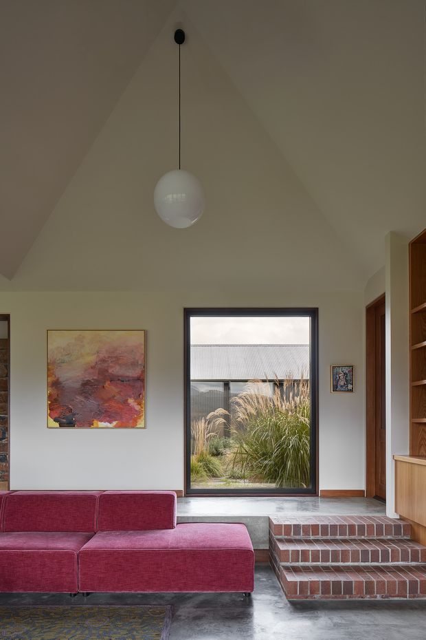 A refined living room with a sculptural vaulted ceiling, white globe pendant light, large dark-framed window, built-in timber shelving, and a vivid burgundy sectional sofa. The room combines polished concrete flooring, exposed brick steps, warm timber joinery, and colourful abstract artwork. Grasses and a neighbouring corrugated roof are visible through the window.