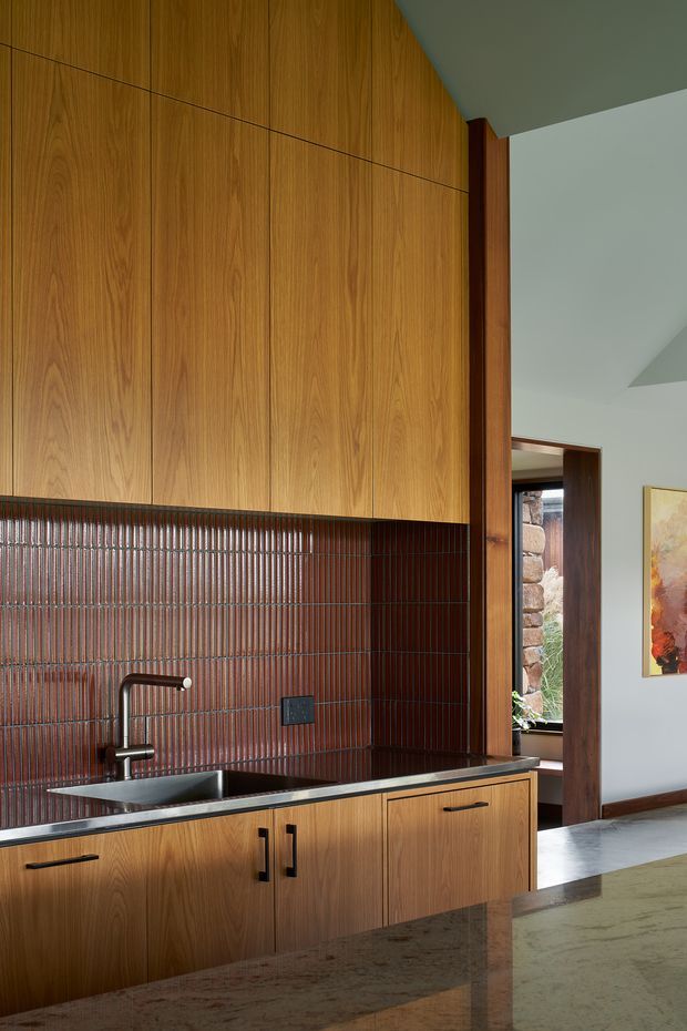 A refined contemporary kitchen featuring extensive warm wood-grain cabinetry, a dark stone benchtop, an integrated stainless-steel sink and tap, and a vertically ribbed dark red-brown tiled splashback. A timber-framed opening leads to an exterior stone wall, while pale walls and a polished stone island surface complete the space.