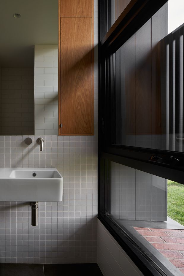 A compact contemporary bathroom featuring a wall-mounted white ceramic basin, chrome wall-mounted tapware, pale square tiled walls, a tall timber vanity cabinet, and large black-framed windows. Strong sunlight creates dramatic shadows across the tiled surfaces, with an exterior brick terrace and lawn visible beyond the glazing.