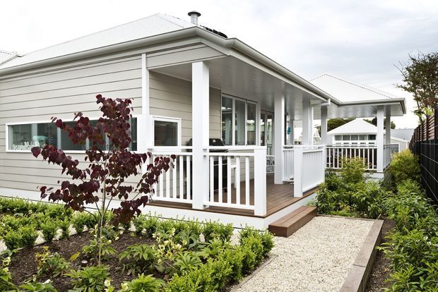 Exterior view of a light-coloured single-storey home with a broad pitched roof, weatherboard cladding, white trim, and a covered raised veranda with white balustrades. A landscaped garden and pale gravel path lead to the entry, with another residential building visible beyond.