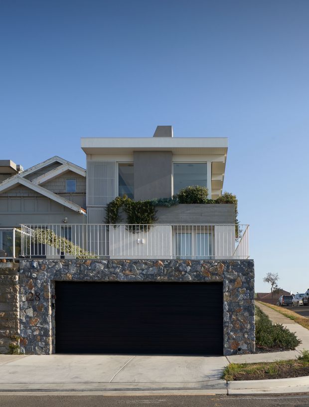 Front exterior view of a contemporary multi-level residence on a sloping suburban site. The upper storey has a flat roof, pale rendered walls, large glazed openings, vertical screening and a planted terrace behind a white metal balustrade. A dark double garage is integrated into a lower level clad in irregular natural stone, with neighbouring homes visible on either side.