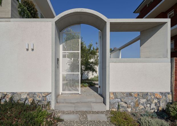 Front entry gate and boundary wall of a contemporary residence, featuring a rendered concrete wall, arched pedestrian opening with a tall white metal slatted gate, and a lower base clad in irregular natural stone. Gravel paving and low landscaping lead to the entrance under a clear blue sky.