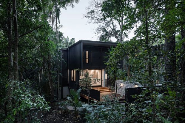 A dark, contemporary two-storey dwelling nestled within dense tropical vegetation. The compact rectangular structure features dark vertical cladding, large screened or glazed openings, a recessed upper-level balcony or void, and a warm timber deck and illuminated ground-floor living area. The architecture is partially concealed by lush forest planting.