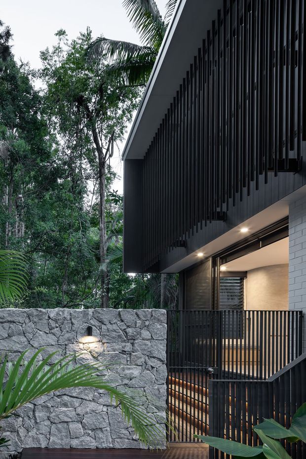 A contemporary multi-level house set within dense tropical vegetation. The dark facade features extensive vertical metal screening and projecting slatted elements, with a deep roof overhang and warm recessed lighting. A natural rough-stone retaining wall, black metal balcony railings, timber decking, and an illuminated sheltered entry create a strong contrast between the dark architecture and lush greenery.