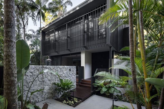 A contemporary dark-clad multi-level house set within lush tropical landscaping. The facade features extensive vertical black metal screening, dark wall cladding, a recessed entry, timber steps, and a low rough-stone boundary wall. Dense palms and broad-leaf planting surround the concrete path and create a private garden setting.