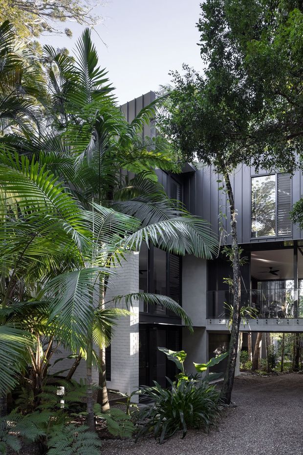 A contemporary multi-level residential building nestled within dense tropical landscaping. The dark grey and white facade features vertical metal cladding, large glazed openings, louvred screens, and elevated structural elements, with a gravel pathway and lush palms surrounding the architecture.
